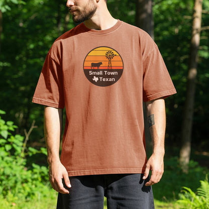 person standing outdoors wearing a burnt orange t-shirt with a circular graphic showing a sunset, windmill, and cow silhouette, with the text Small Town Texan and a small outline of the state of Texas.